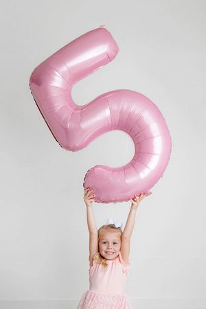 Happy little blonde girl in pink dress lifting a large pink number 5 balloon overhead.
