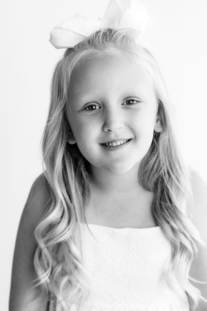 Smiling little girl with long blonde wavy hair and white bow, wearing a white dress in a black and white portrait by Temecula photographer.