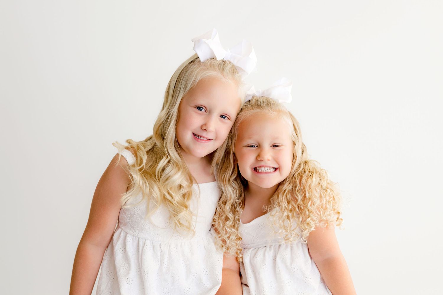 Two young blonde girls in white dresses and white bow hair accessories smiling together against a white background.