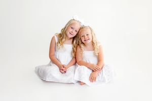 Two young girls in matching white dresses with curly blonde hair smile together against a white background.