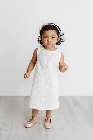 Toddler girl in white textured dress with headband and pink shoes on light background.