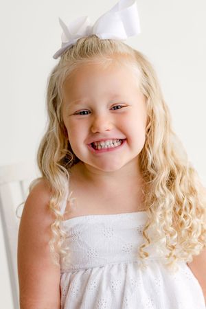 Smiling young blonde girl with curly hair wearing a white dress and bow, portrait photo on light background.