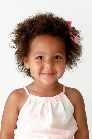 Adorable toddler girl with curly hair and a pink bow smiles in a light studio portrait.