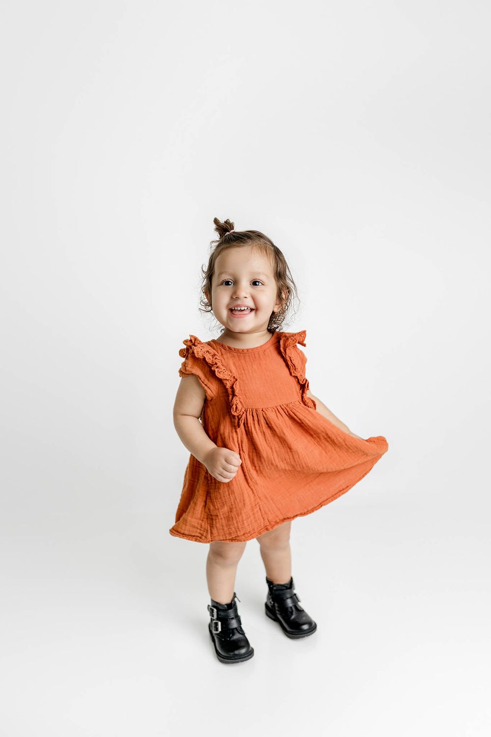 Happy toddler girl in rust orange ruffled dress and black boots twirling and smiling.