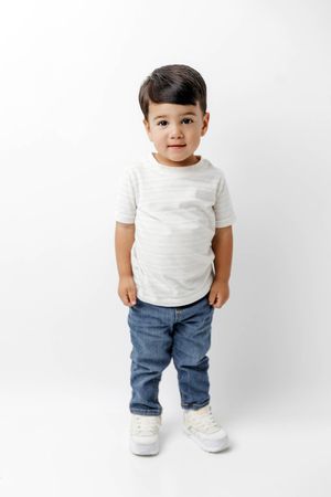 Cute toddler boy in a striped tee and jeans standing against a white studio background.