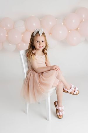 Little girl in a blush tulle dress sitting on a white chair surrounded by pink balloon garland decor.