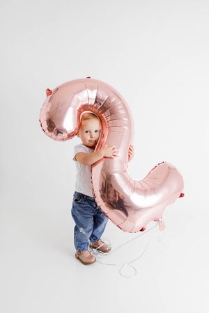 Toddler child in jeans and white tee hugging a large rose gold number 2 balloon on white background.