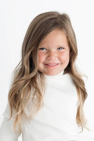 Smiling little girl with long wavy hair wearing a white turtleneck top against a white background.