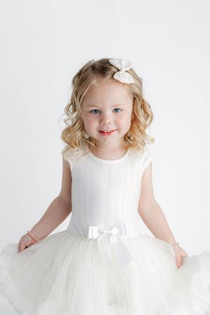Adorable blonde toddler girl in a white tulle dress with a bow hairclip smiling at the camera.