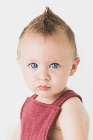Curious baby with blue eyes and a mohawk hairstyle wearing a red tank top against white background.