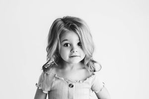 Black and white portrait of a little girl with wavy blonde hair in a ruffled top on white background.