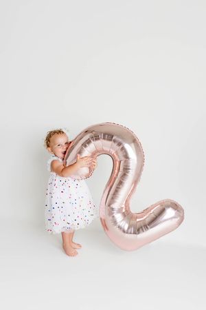 Toddler girl in white polka dot dress hugging a rose gold number 2 balloon on white background.