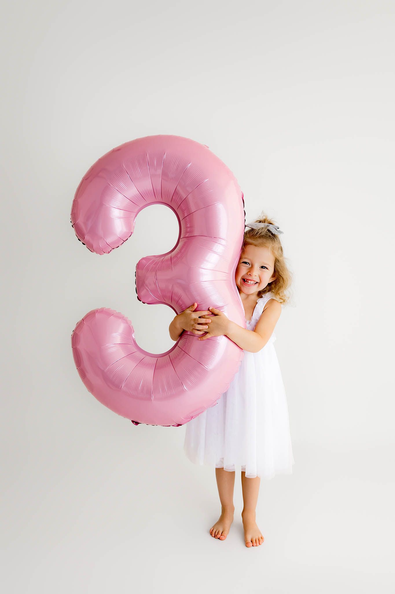 A smiling little girl in a white dress holds a large pink number 3 balloon, celebrating her 3rd birthday.
