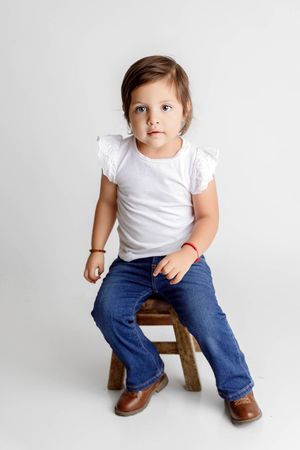 Toddler girl in white ruffle top and jeans sitting on wooden stool against white background.