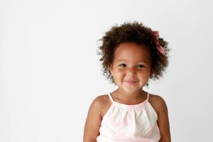 Smiling little girl with curly hair and pink bow wearing a light pink dress.