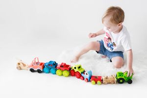 Baby boy in white shirt and denim shorts playing with colorful toy vehicles on white fur rug.
