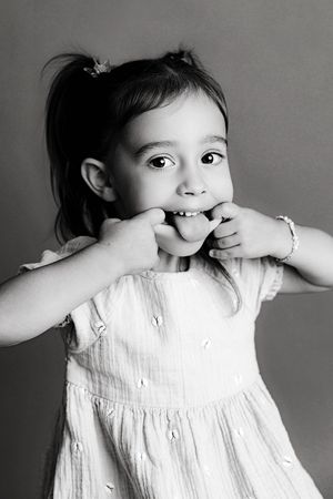 3rd birthday photographer in Menifee: Black and white portrait of a toddler girl in a white dress playfully covering her mouth with hands.