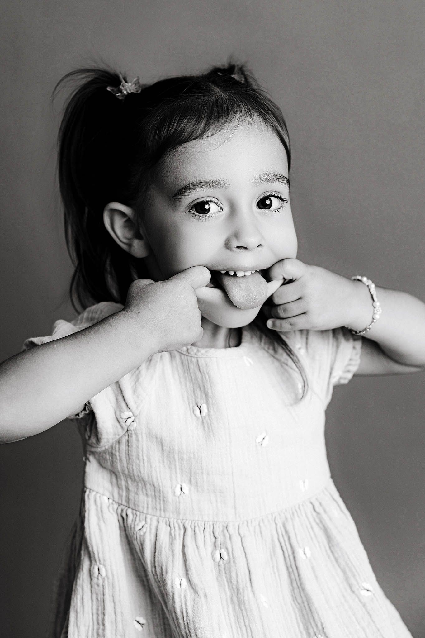 3rd birthday photographer in Menifee: Black and white portrait of a toddler girl in a white dress playfully covering her mouth with hands.