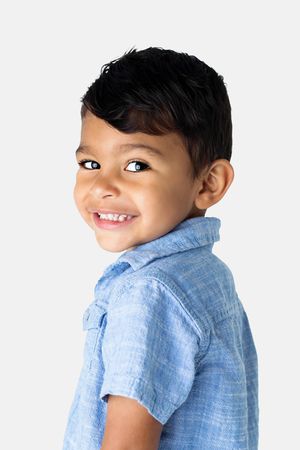 Adorable young boy in a blue shirt smiles over his shoulder in a studio portrait photo.