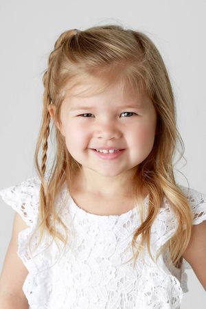 Smiling blonde girl in a white lace dress poses for a bright studio portrait photograph.