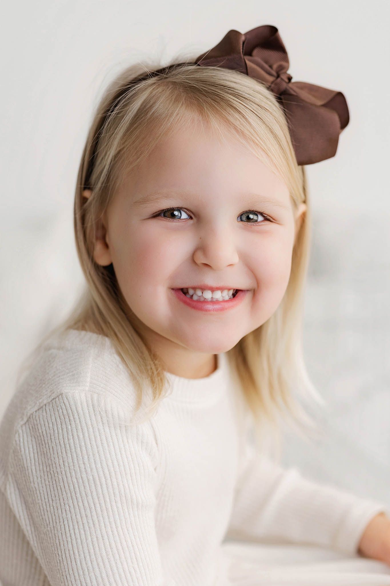 Cheerful blonde little girl with a brown bow headband smiles in a bright white studio portrait.
