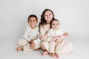 Three siblings in cream outfits sit together on a white background in Menifee kids photography studio.