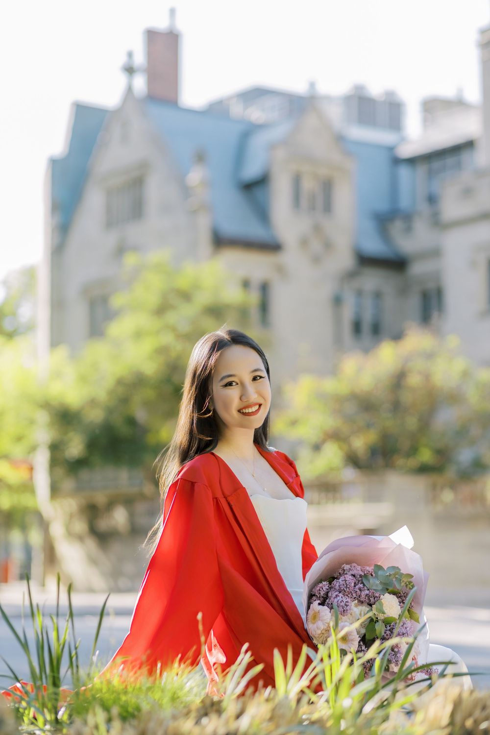 Boston University graduate holding flower bouquet, smiling outdoors near historic stone building on sunny day.