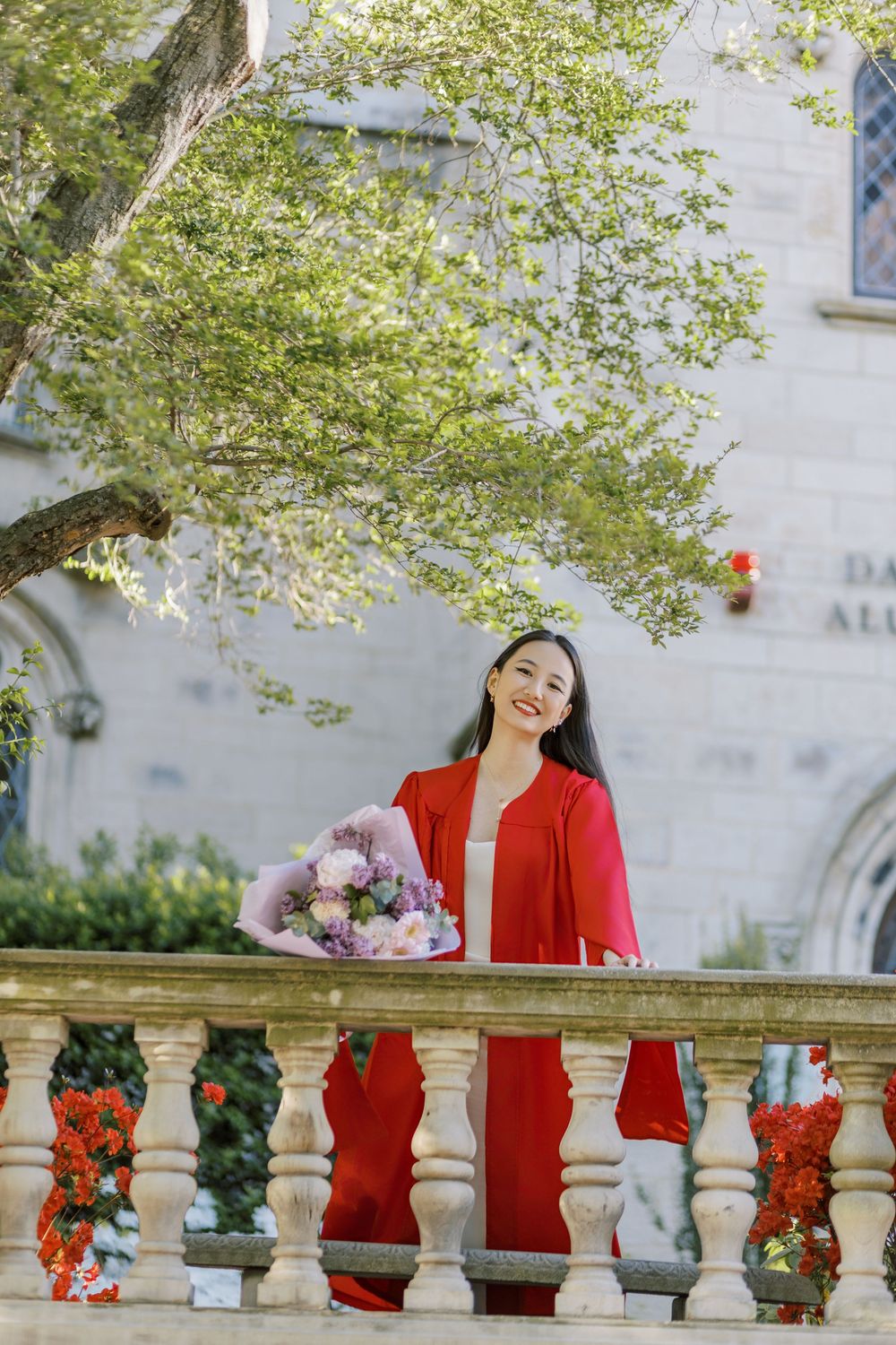 A Boston University graduate holding a floral bouquet beside a stone balustrade, surrounded by lush greenery.