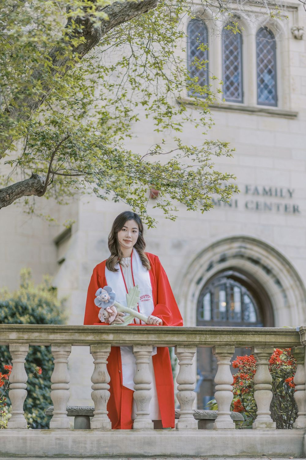 Boston University graduate in red graduation gown holding flowers stands by stone balustrade at university alumni center.