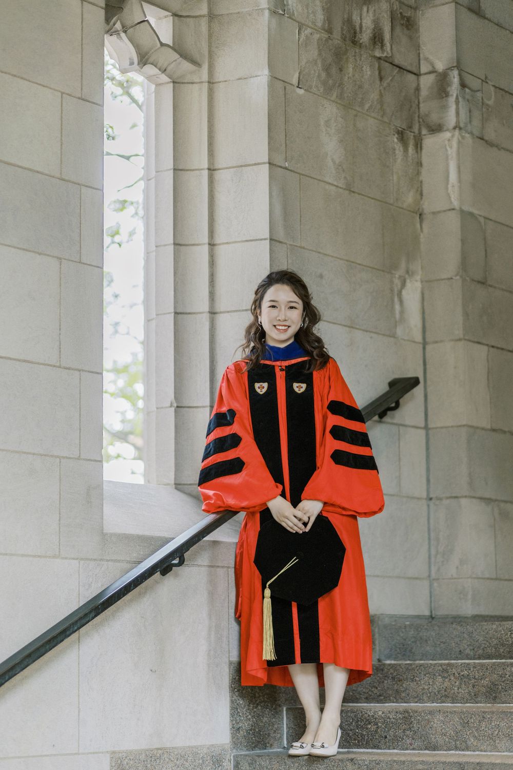 A Boston University graduate in red doctoral regalia stands on stone steps holding a cap at a university building.