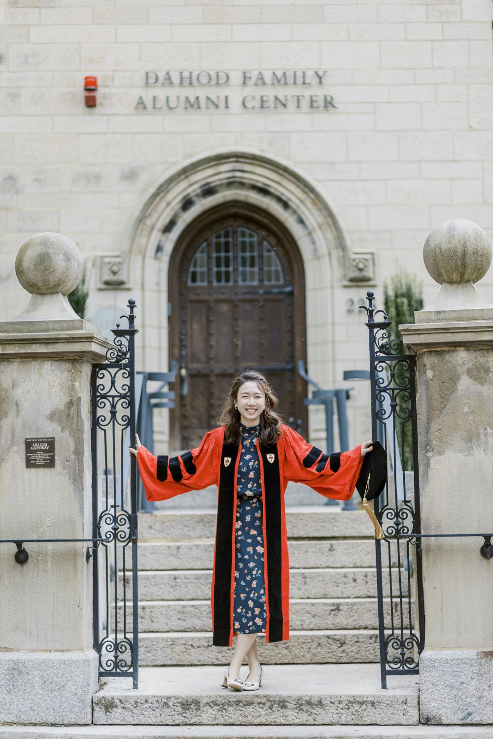 Boston University Graduate in red doctoral robes celebrates at Dahod Family Alumni Center steps, arms wide open in joy.