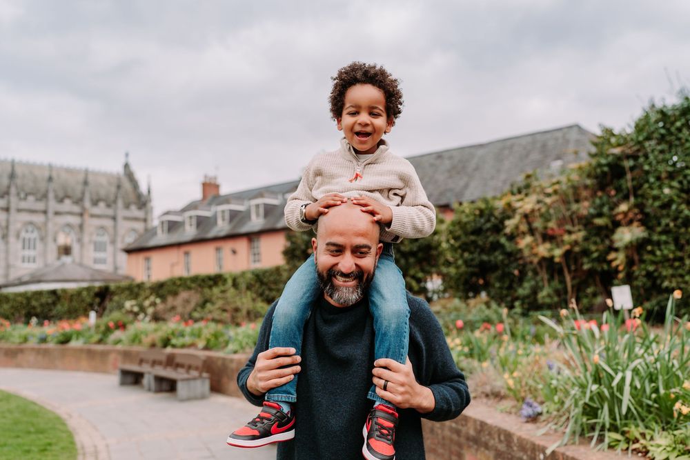 A joyful child sits on a smiling man's shoulders in a lush garden, both laughing outdoors near a historic building.