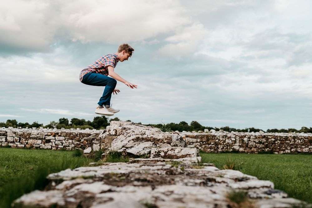 Young boy jumping between ancient stone walls on a cloudy day in a green countryside landscape.