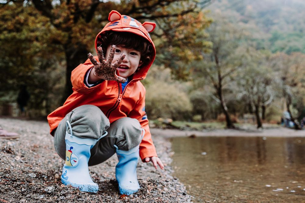 A happy toddler in an orange bear hoodie and blue rain boots crouches by a rocky riverbank in autumn.