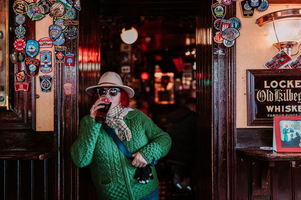 Person in green jacket and hat drinking outside a vintage pub with whiskey signage and decorative badges.