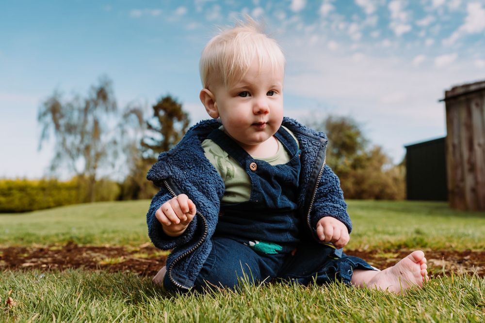 A cute baby in a blue jacket sits on green grass outdoors under a blue sky.