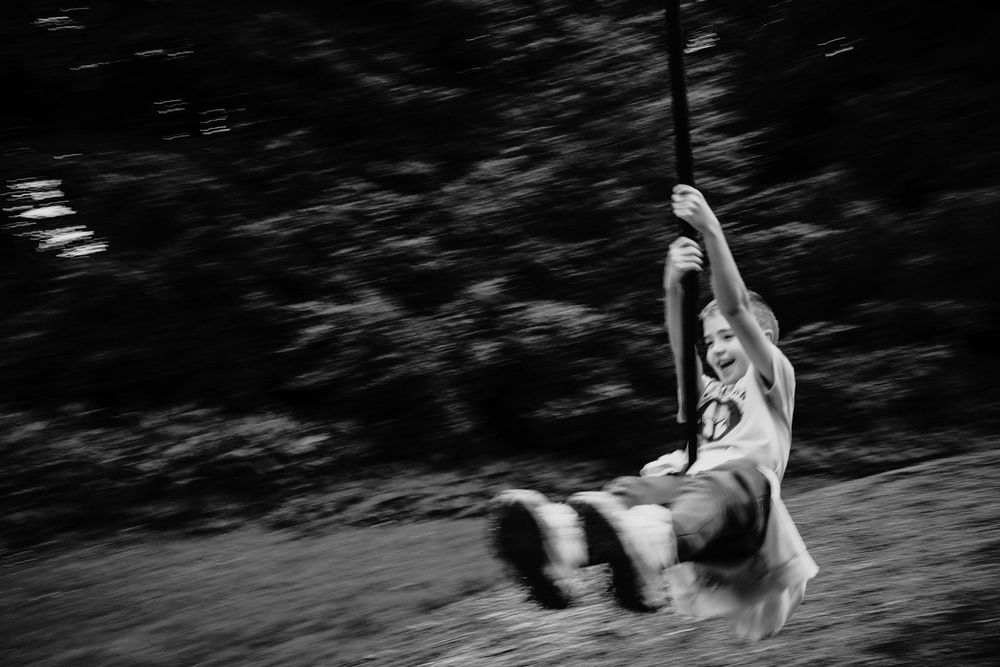 Black and white photo of a child joyfully riding a zip line through a blurred forest background.