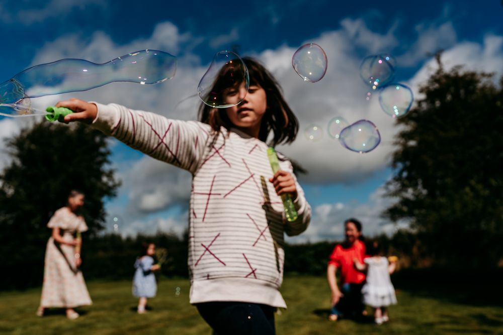 A young girl blows bubbles outdoors in a park on a cloudy day, with children playing in the background.