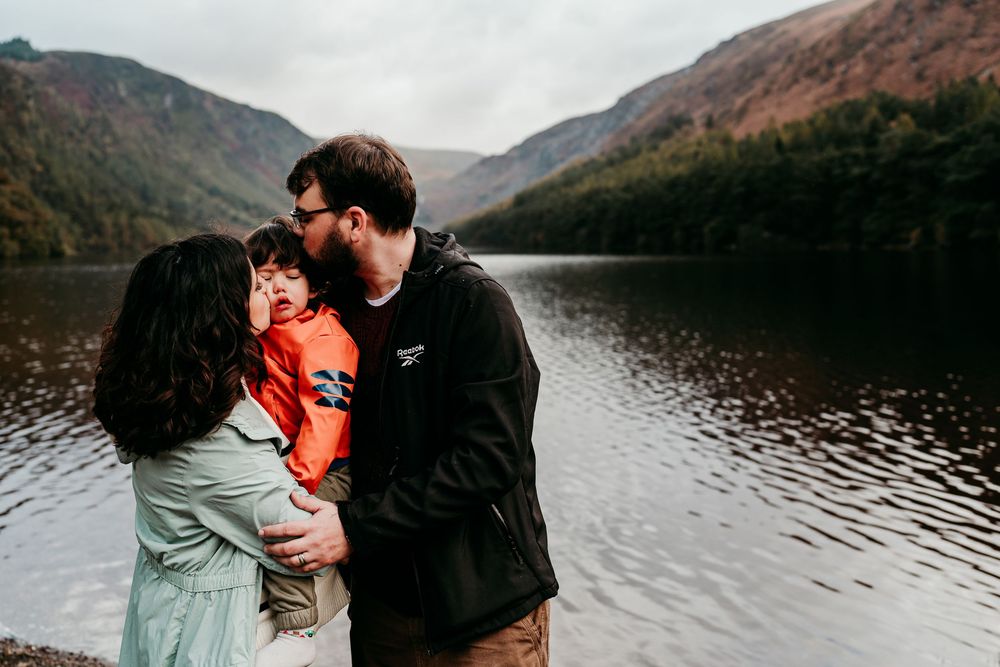 Family of three sharing a tender moment by a scenic mountain lake with lush autumn hills in the background.