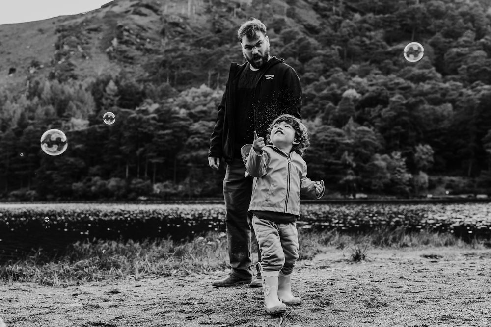 Black and white photo of a father and toddler playing with bubbles outdoors near a lake and hills.