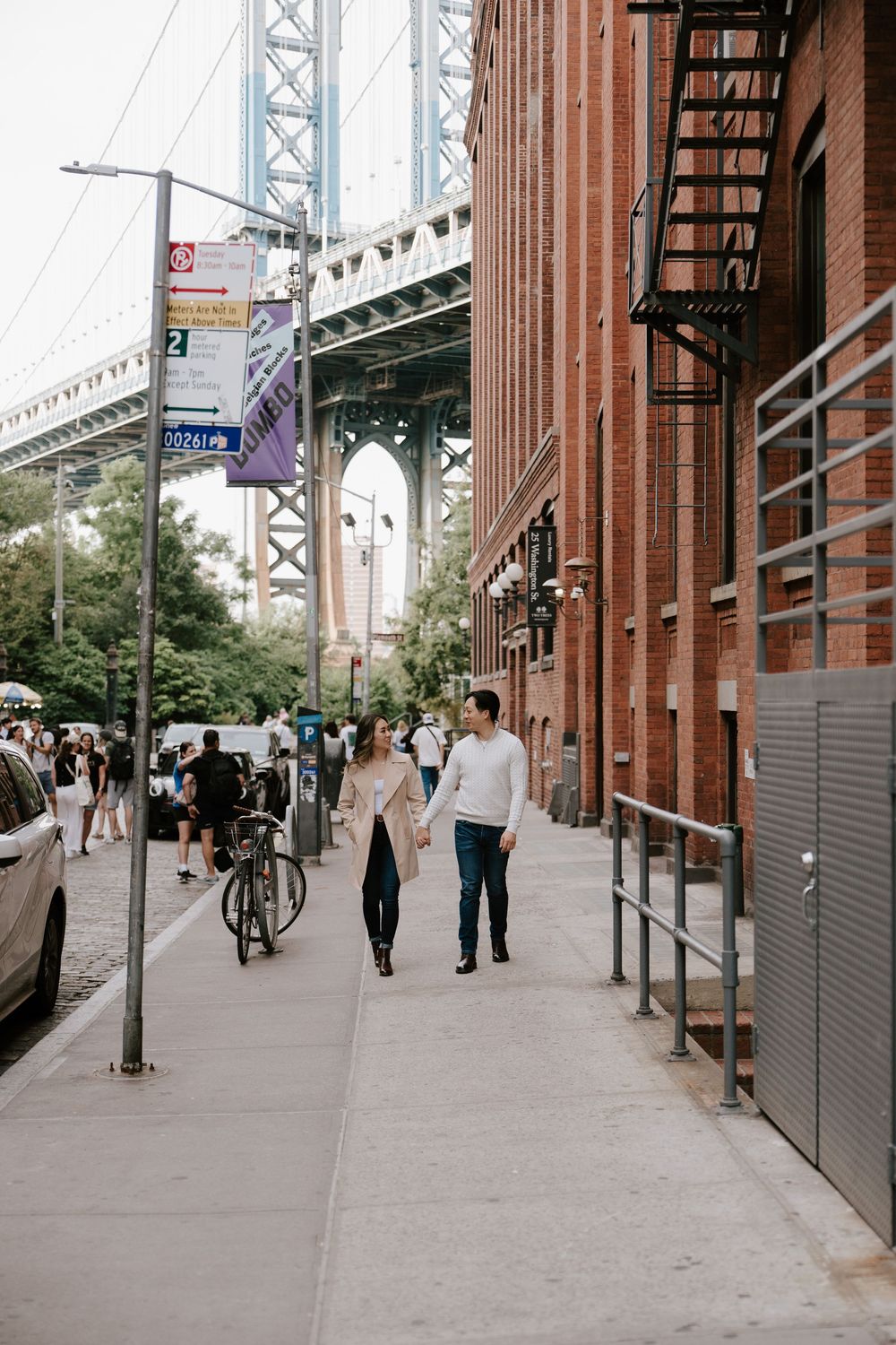 Couple holding hands walking on a Brooklyn sidewalk near the Manhattan Bridge and a red brick building.