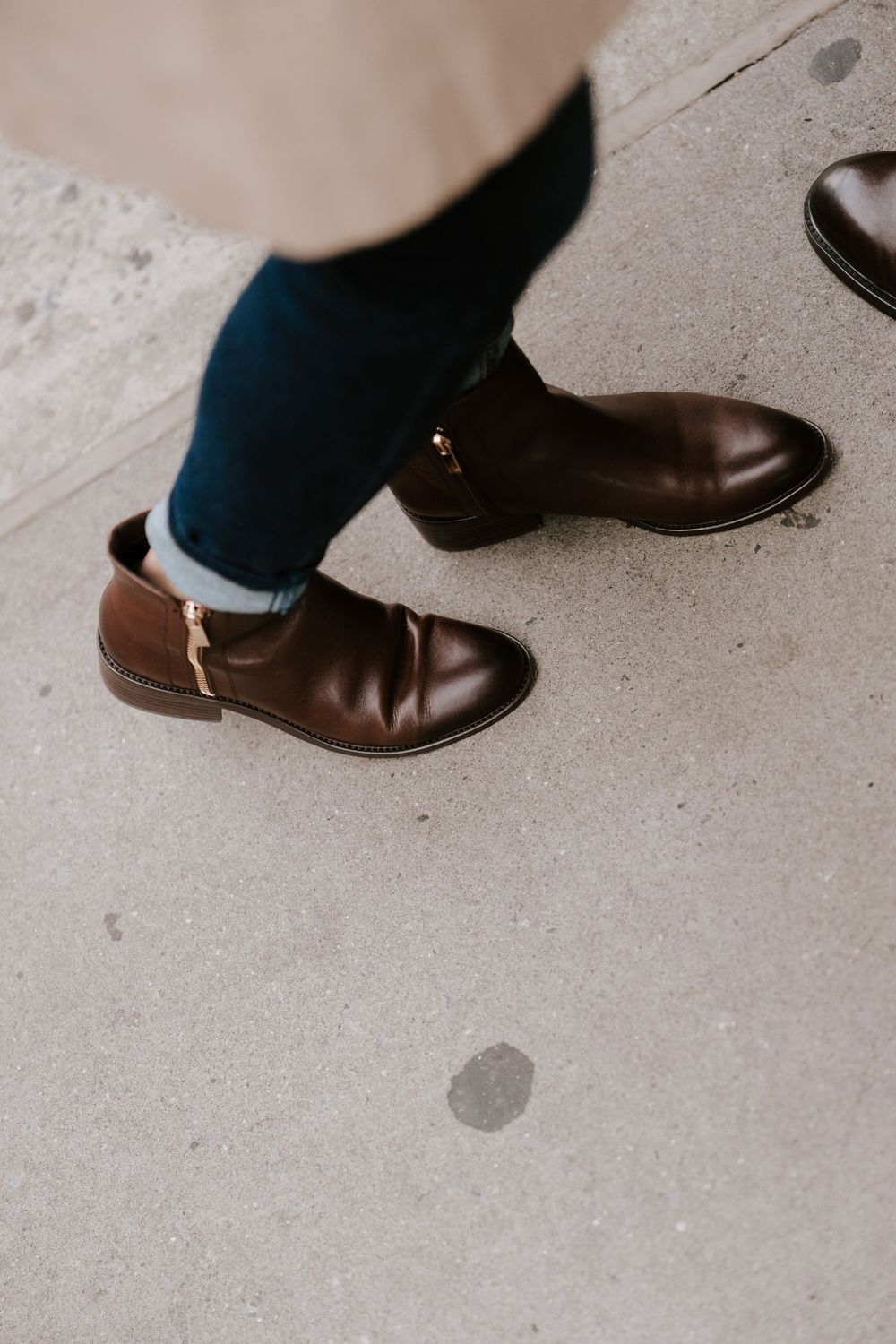 Brown leather ankle boots with gold zipper detail, paired with blue jeans on a concrete surface.