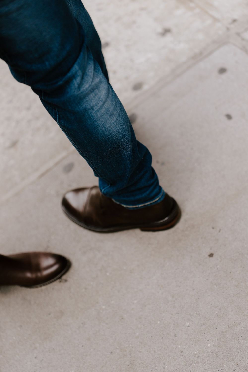 Close-up of a person wearing blue jeans and brown leather dress shoes walking on a concrete sidewalk.