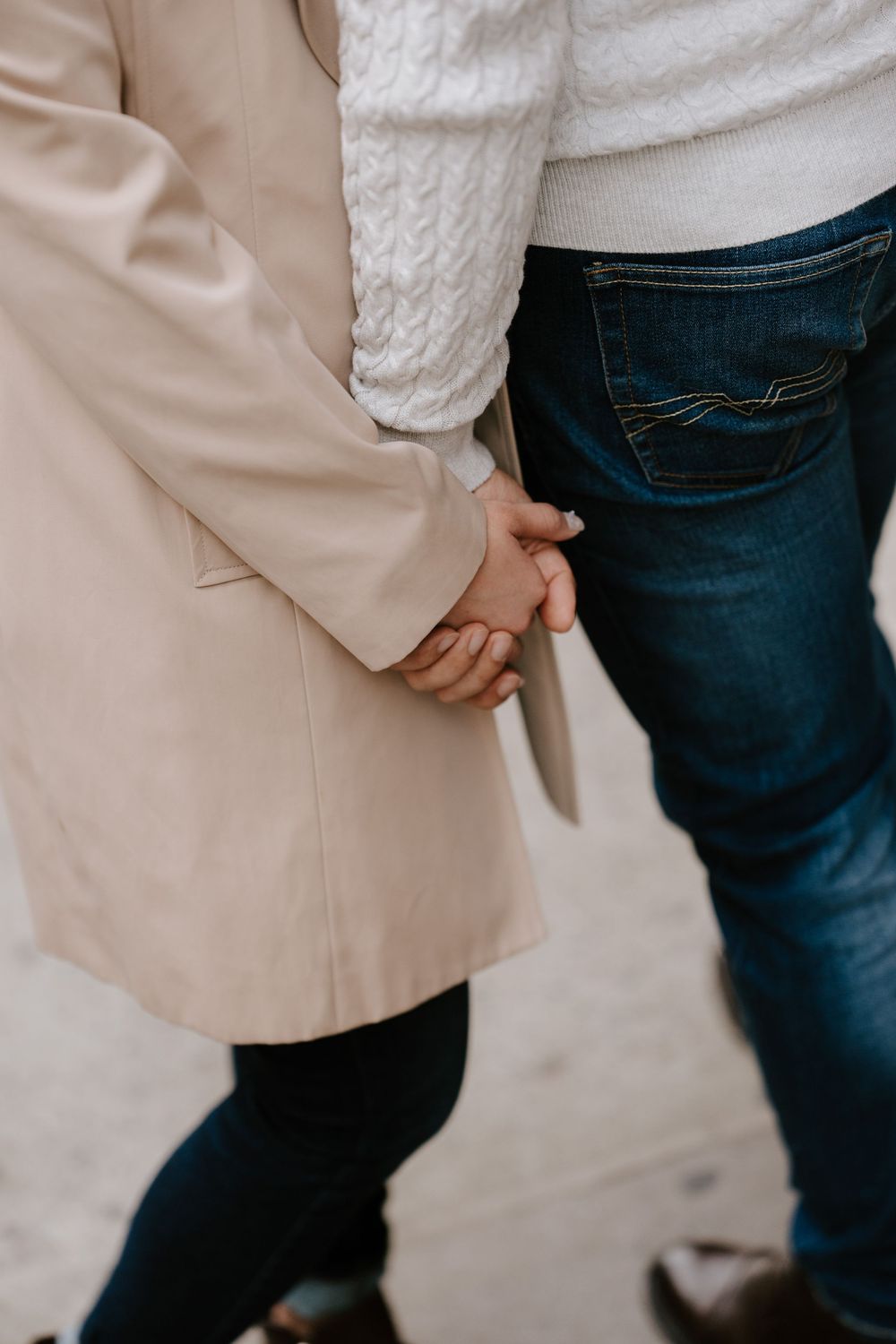 Couple holding hands outdoors, woman in beige trench coat and man in dark jeans and white sweater.