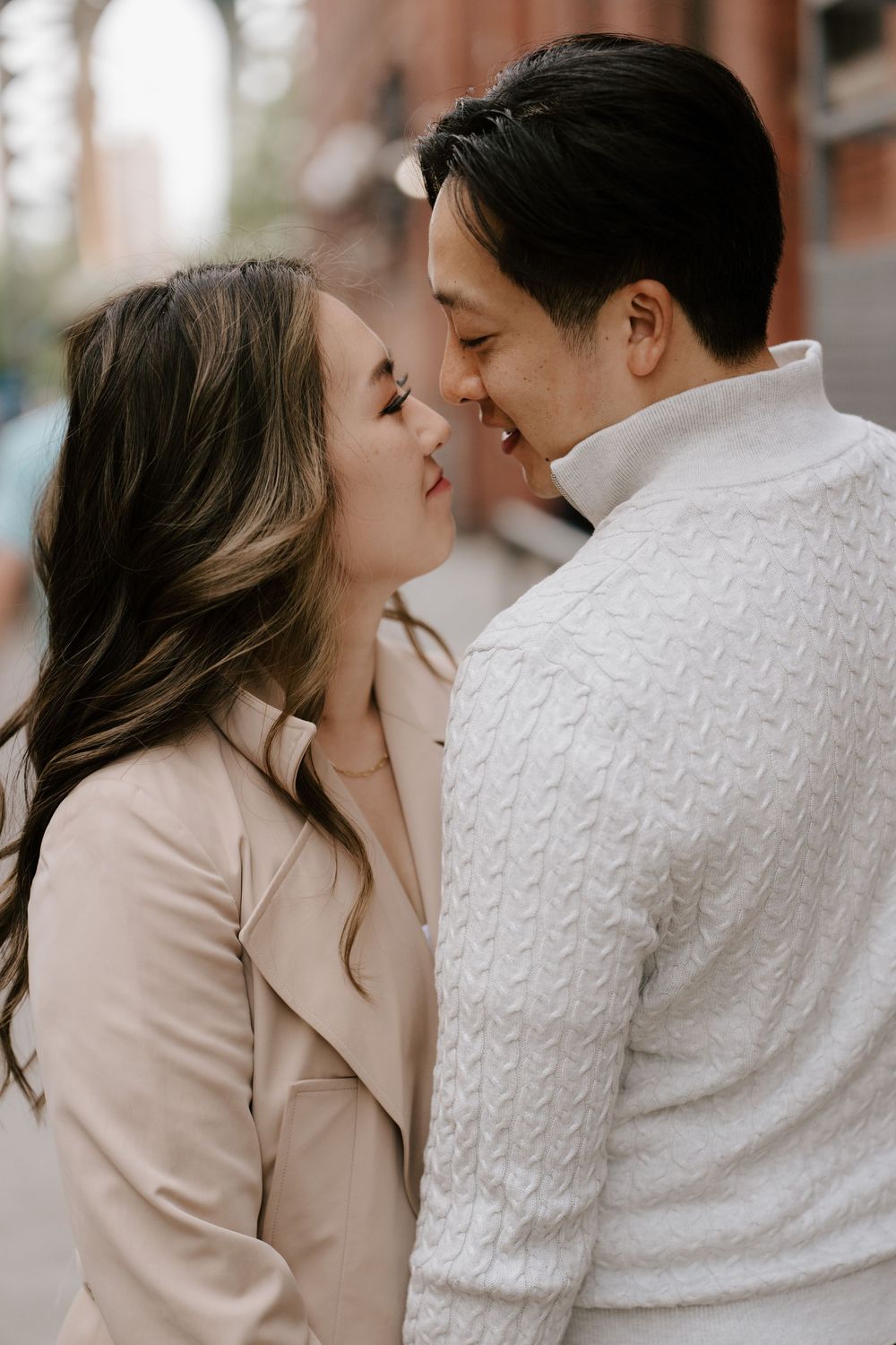 A couple shares a romantic almost-kiss moment on a city street, dressed in elegant neutral tones.