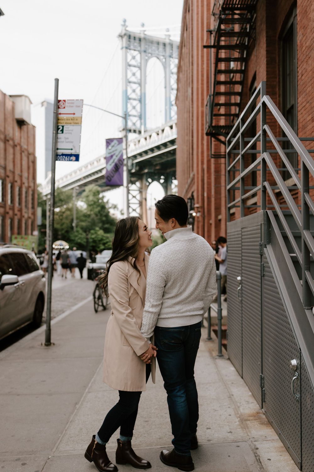 Couple holding hands on a Brooklyn sidewalk with the Manhattan Bridge in the background on a cloudy day.