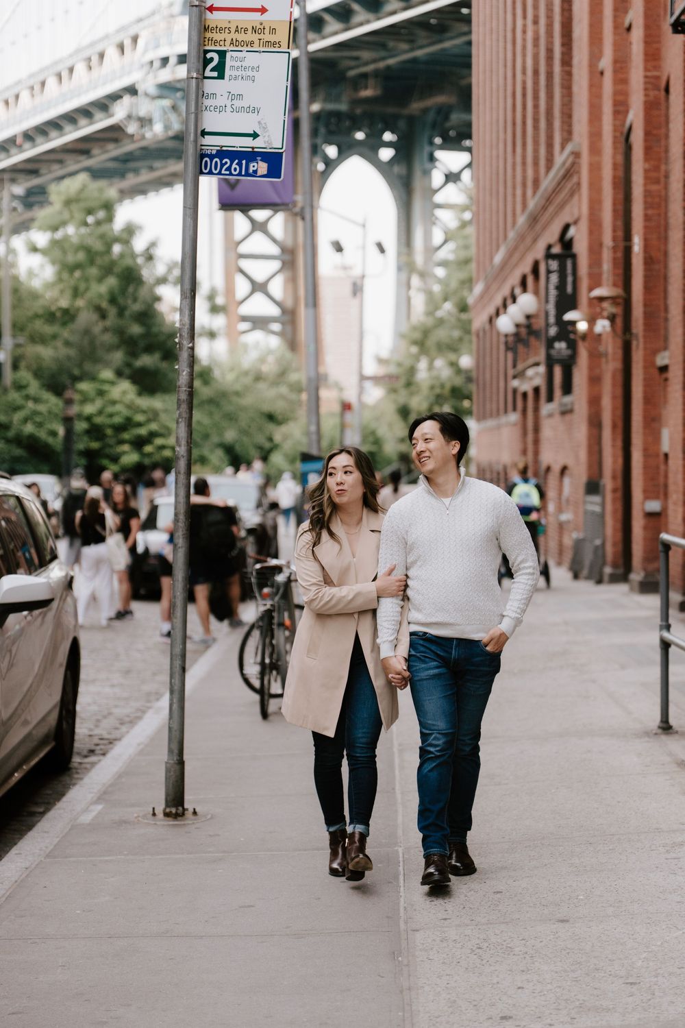 Couple walking hand in hand on a Brooklyn sidewalk near Manhattan Bridge, woman in pink coat and man in white sweater.