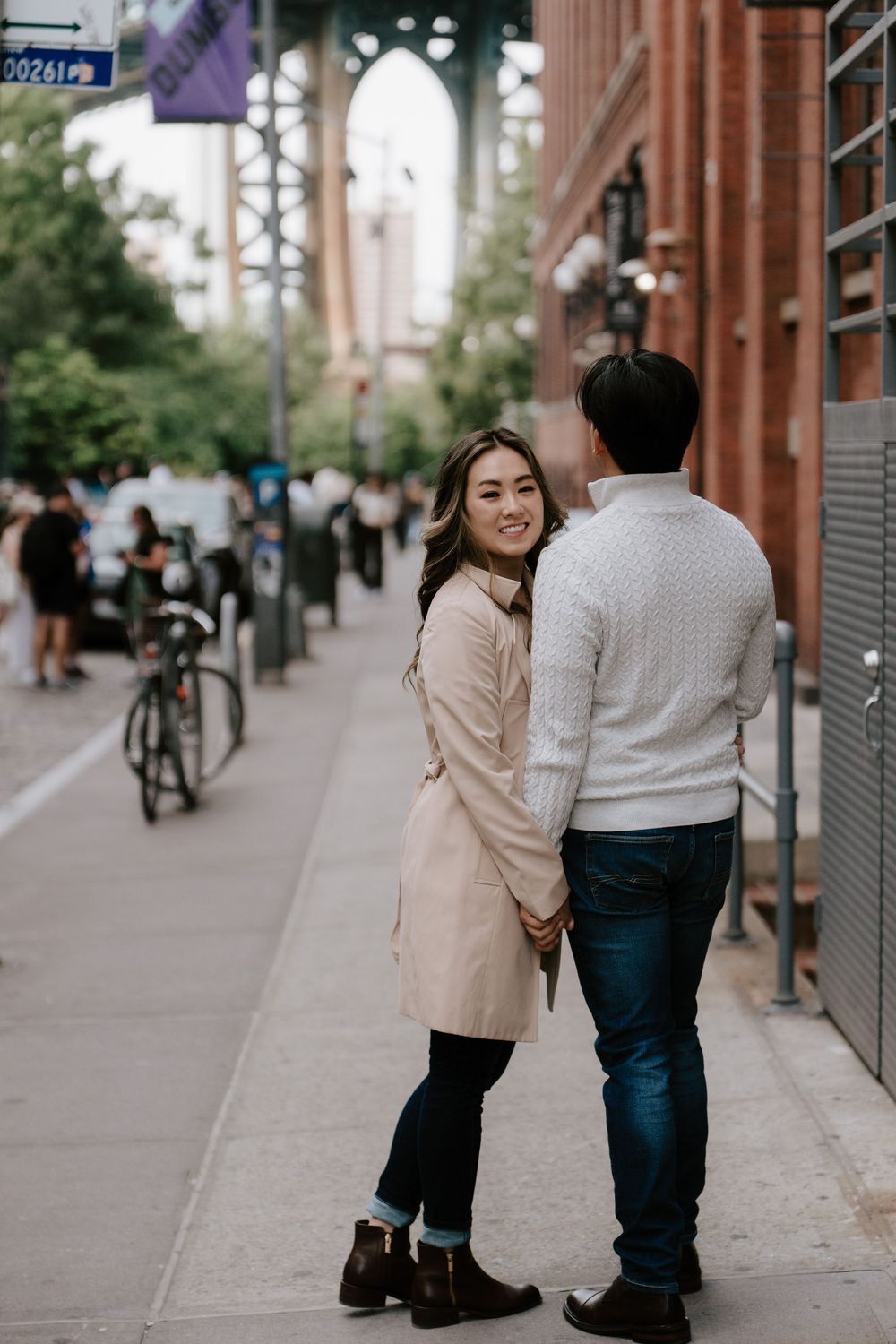 Couple holding hands on Brooklyn sidewalk, woman in pink trench coat smiling back at camera near Manhattan Bridge.