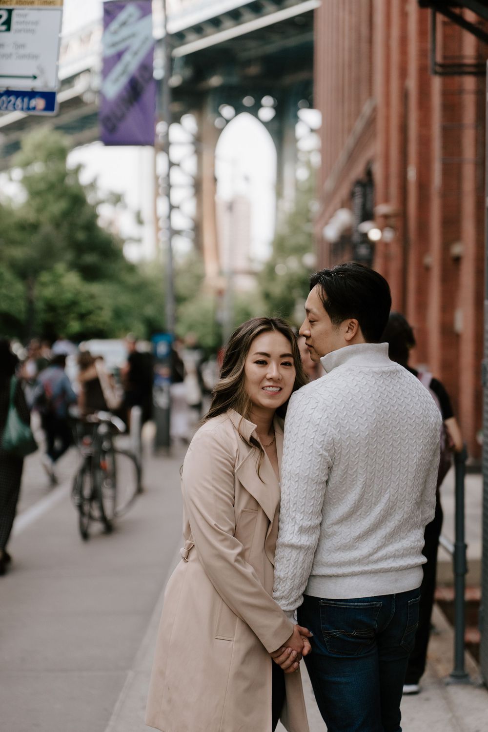 Smiling Asian couple embracing on urban Brooklyn sidewalk with Manhattan Bridge arch visible in background.