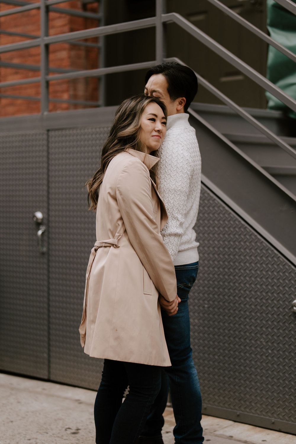 A couple embraces on a city street, woman in beige trench coat smiling over shoulder, man in white sweater and jeans.